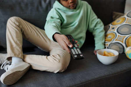 Cropped shot of little boy in hoodie and pants choosing tv channel while sitting on couch in living room and pressing buttons on remote controlの写真素材