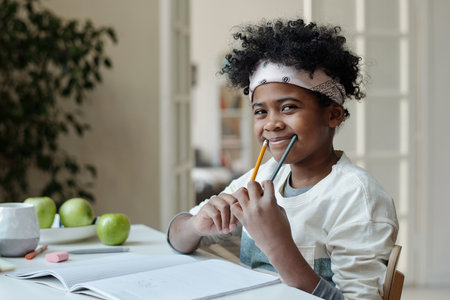 Cute African American schoolboy having fun while sitting by desk in living room and holding two pencils by edges of mouth and making smileの写真素材