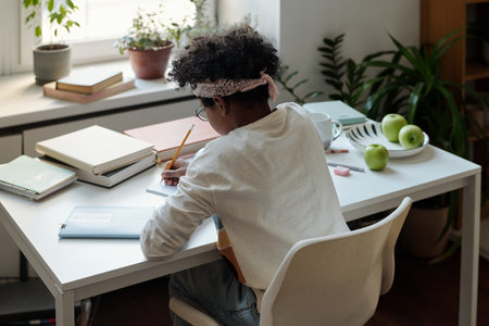 Back view of diligent African American schoolchild in white pullover writing essay or making notes...
