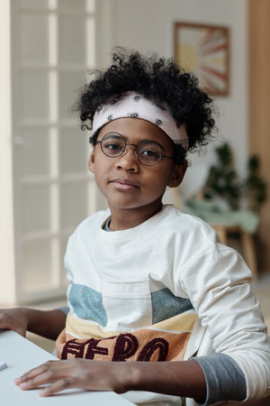 Adorable African American schoolchild in headband, casual pullover and eyeglasses with round frame...