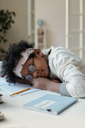 Cute schoolboy napping by desk after preparing home assignment while keeping head on open copybook...