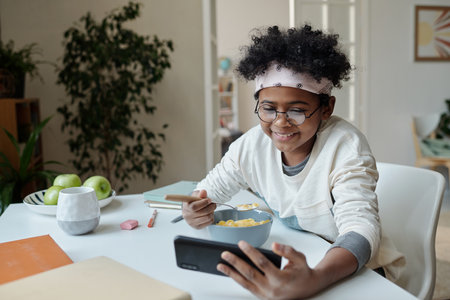 Smiling schoolchild looking at smartphone screen and eating muesli with milk for breakfast while...