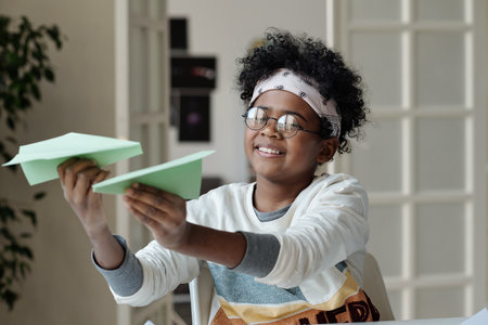Happy African American child in casualwear and eyeglasses playing with handmade paper planes in front of camera in living roomの写真素材