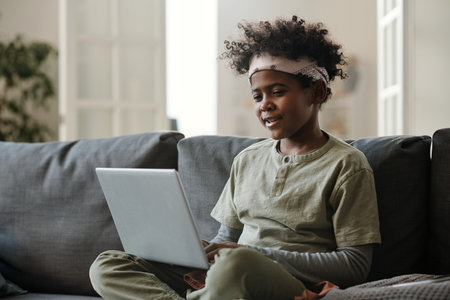 African American boy in pajamas and headband sitting on couch and looking at laptop screen while...