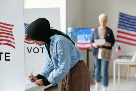 Young Muslim woman in hijab bending over vote booth and making her choice of candidate during presidential elections at polling placeの写真素材