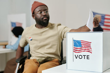 Box for ballot papers on desk and young African American man with disability sitting in wheelchair and making his choiceの写真素材