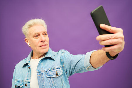 Confident aged man with grey hair holding smartphone in hand while stretching arm during online communication or taking selfieの写真素材