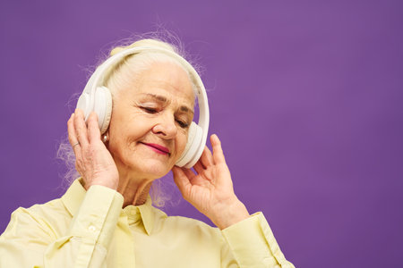 Smiling senior woman with white hair touching headphones while standiing against lavender background and enjoying her favorite musicの写真素材