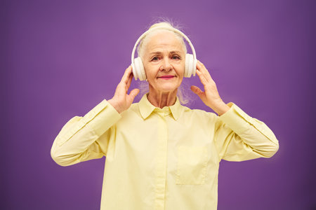 Happy aged woman in yellow shirt listening to music in headphones and looking at camera with smile...