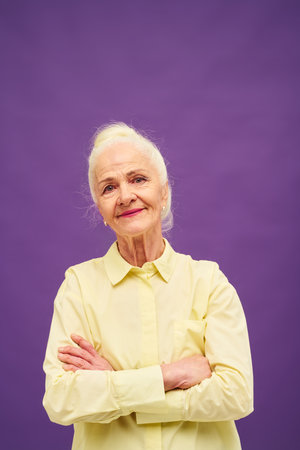 Happy female representative of silver society in yellow shirt looking at camera while standing in isolation and keeping arms crossed by chestの写真素材