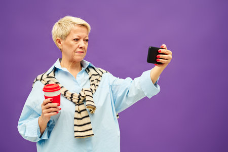Mature blond woman in casualwear having coffee and watching online video while holding smartphone in front of herself in isolationの写真素材