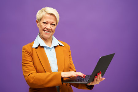 Happy mature businesswoman in formalwear looking at camera and looking through online data in laptop while standing in isolationの写真素材