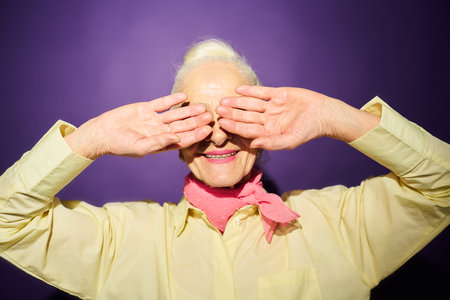 Smiling senior woman in yellow shirt covering eyes by her hands while hiding from bright light while standing against lavender backgroundの写真素材