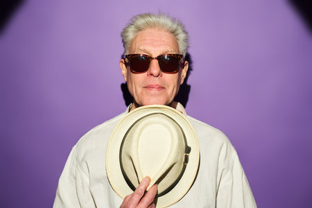 Serious mature man with grey hair holding white panama hat by his chest while standing in front of camera over violet backgroundの写真素材