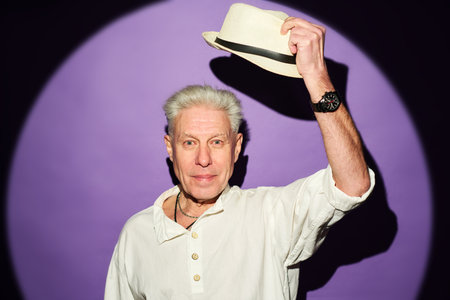 Mature man with white hair taking off his panama hat in front of camera while standing against lit lavender background in isolationの写真素材