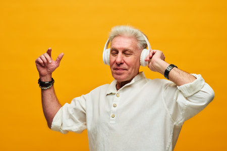 Happy aged man in white shirt and headphones enjoying music while standing in front of camera in isolation against yellow backgroundの写真素材