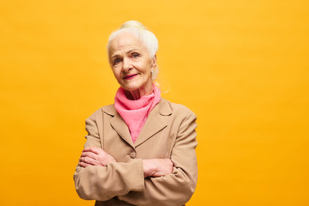 Senior woman in beige blazer and pink scarf crossing her arms by chest and looking at camera while standing in isolation over yellow backgroundの写真素材