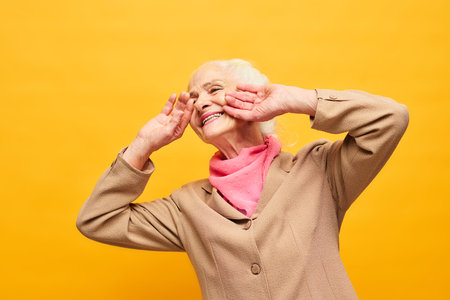 Joyful senior woman in beige blazer and pink scarf touching face by hands while laughing in front of camera against yellow backgroundの写真素材