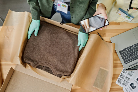 Top view of gloved hands of volunteer putting folded woolen knitted sweater into box and packing it...