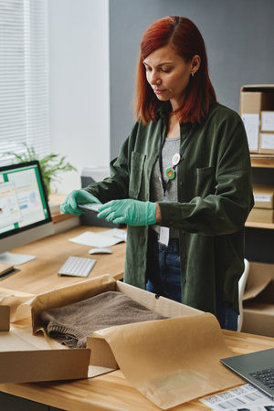 Young female volunteer with smartphone taking photo of folded sweater in open box before packing it and sending to people in needの写真素材