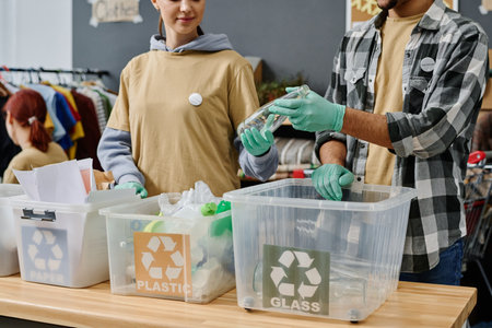 Young woman in gloves passing empty bottle to male volunteer while both standing by desk with trash bins and sorting garbageの写真素材