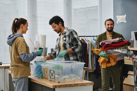 Two young volunteers sorting garbage while man carrying stack of second hand clothes while passing by behind them in volunteering officeの写真素材