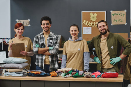 Group of happy young intercultural volunteers looking at camera with smiles while standing by table with stacks of second hand clothesの写真素材