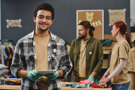 Happy young multi-ethnic male volunteer in casualwear and protective gloves holding tablet while standing against group of colleaguesの写真素材