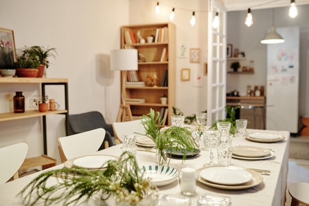 Part of living room with table served for family dinner with plates, wineglasses for drinks, green plants and candles used as decorationsの写真素材