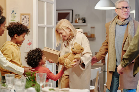 Happy mature blond woman in beige coat passing teddybear to cute little grandson standing in front...