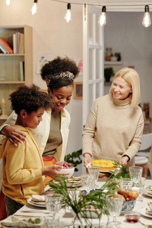 Cute little African American boy helping his mother and grandmother with serving table for festive dinner to celebrate family life eventの写真素材