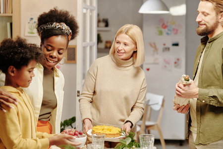 Young smiling woman embracing her cute little son helping her serve table for family dinner among his father and grandmother holding pieの写真素材