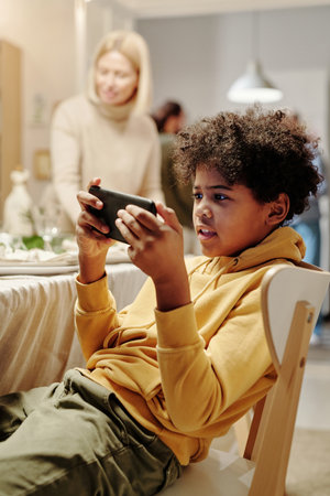 Adorable African American boy watching online video in smartphone while sitting on chair against his grandmother serving table for dinnerの写真素材