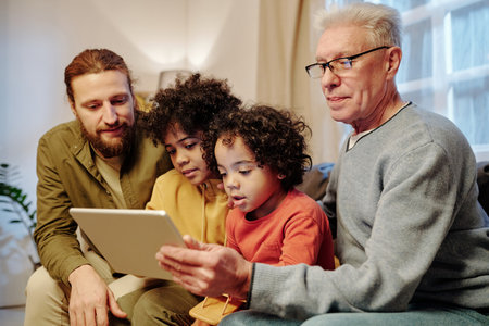 Young and mature men and two cute little boys watching online movie or video in tablet while sitting...