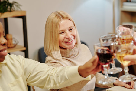 Happy blond mature woman clinking with wineglasses of guests sitting by festive table and enjoying family dinner in honour of life eventの写真素材