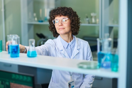 Young female researcher in eyeglasses and lab coat taking test tube containing blue liquid from...