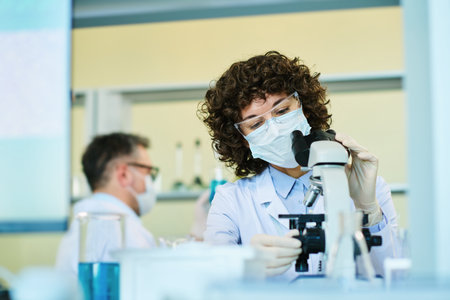 Mature expert in virology wearing protective mask adjusting dial on microscope while studying new...