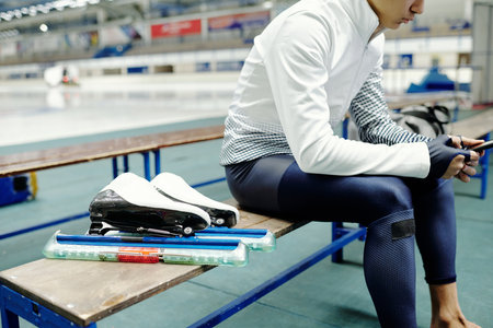Cropped shot of adolescent male ice skater in sports uniform sitting on bench inside arena after training and texting in mobile phoneの写真素材