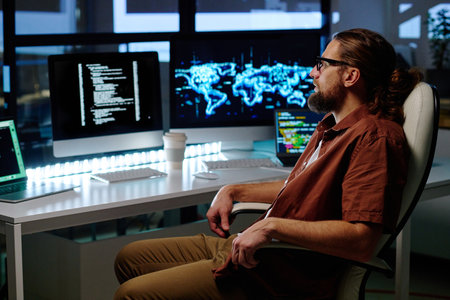 Young serious IT programmer sitting in armchair by workplace in front of several computers with coded data on screens late in the eveningの写真素材