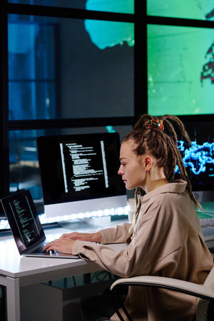 Side view of young female IT support manager with dreadlocks sitting by workplace, looking at laptop screen and typing in dark officeの写真素材