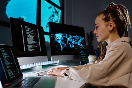 Side view of young programmer developing new software or decoding data on computer screen while sitting by workplace in dark officeの写真素材