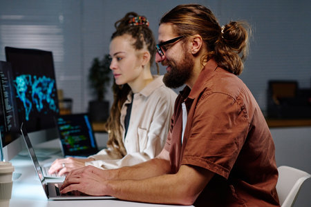 Side view of young male IT engineer typing on laptop keyboard while decoding data on screen against female coworker using computerの写真素材