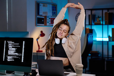 Young tired female programmer with headphones on neck stretching arms over her head while sitting by workplace in front of laptop in officeの写真素材