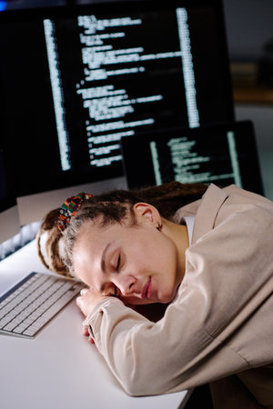 Young tired woman with dreadlocks keeping head on desk by keyboard while sleeping in dark office against computer screens with coded dataの写真素材