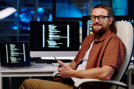 Young smiling male IT engineer in eyeglasses using smartphone and looking at camera while sitting in armchair by his workplaceの写真素材