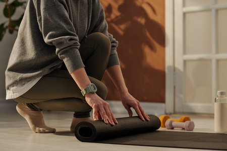 Cropped shot of young active woman in leggins and grey sweatshirt rolling fitness mat on the floor after morning training at homeの写真素材