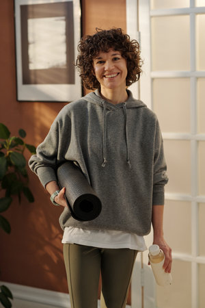 Happy young active woman in grey sportswear holding rolled mat while standing in living room against wall and door and looking at cameraの写真素材