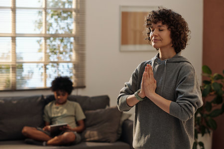 Young relaxed woman in grey hoodie keeping hands put together by chest during yoga exercise while standing in front of camera at homeの写真素材