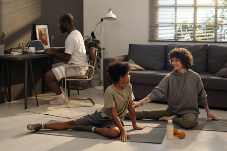 Happy young mother in activewear sitting on mat on the floor of living room and talking to her son during workout against networking manの写真素材