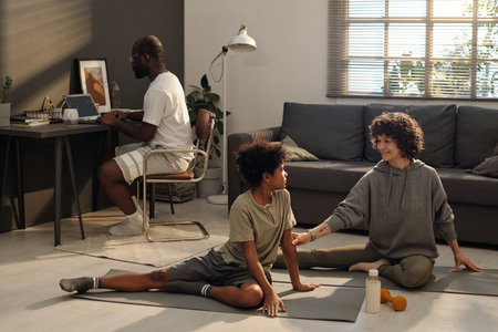 Happy woman helping her little son while both doing stretching exercise on mats on the floor of living room against young man using laptopの写真素材